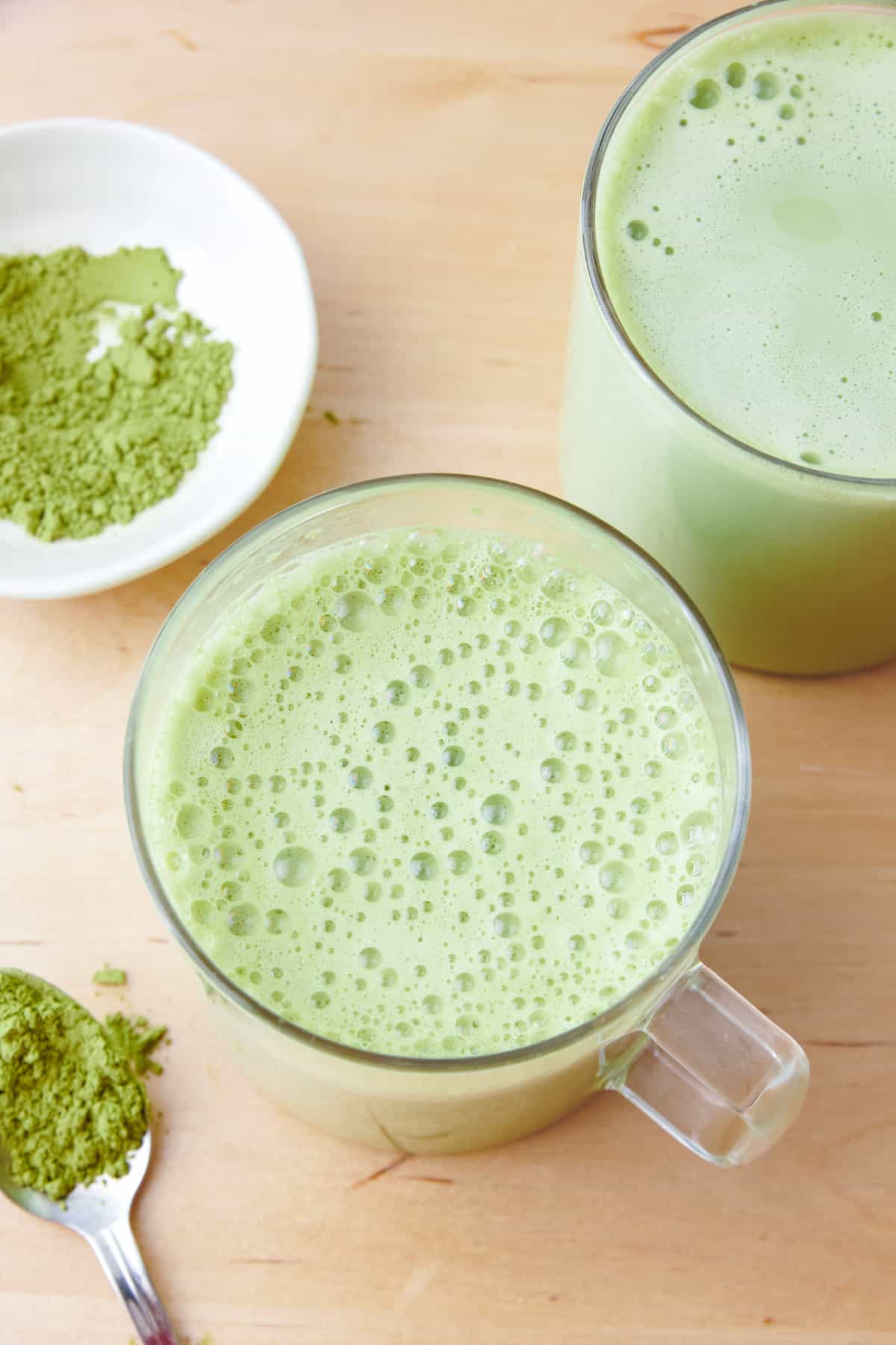 2 glass mugs of matcha latte with focus on one. Spoonful of matcha powder and a small dish of more nearby.