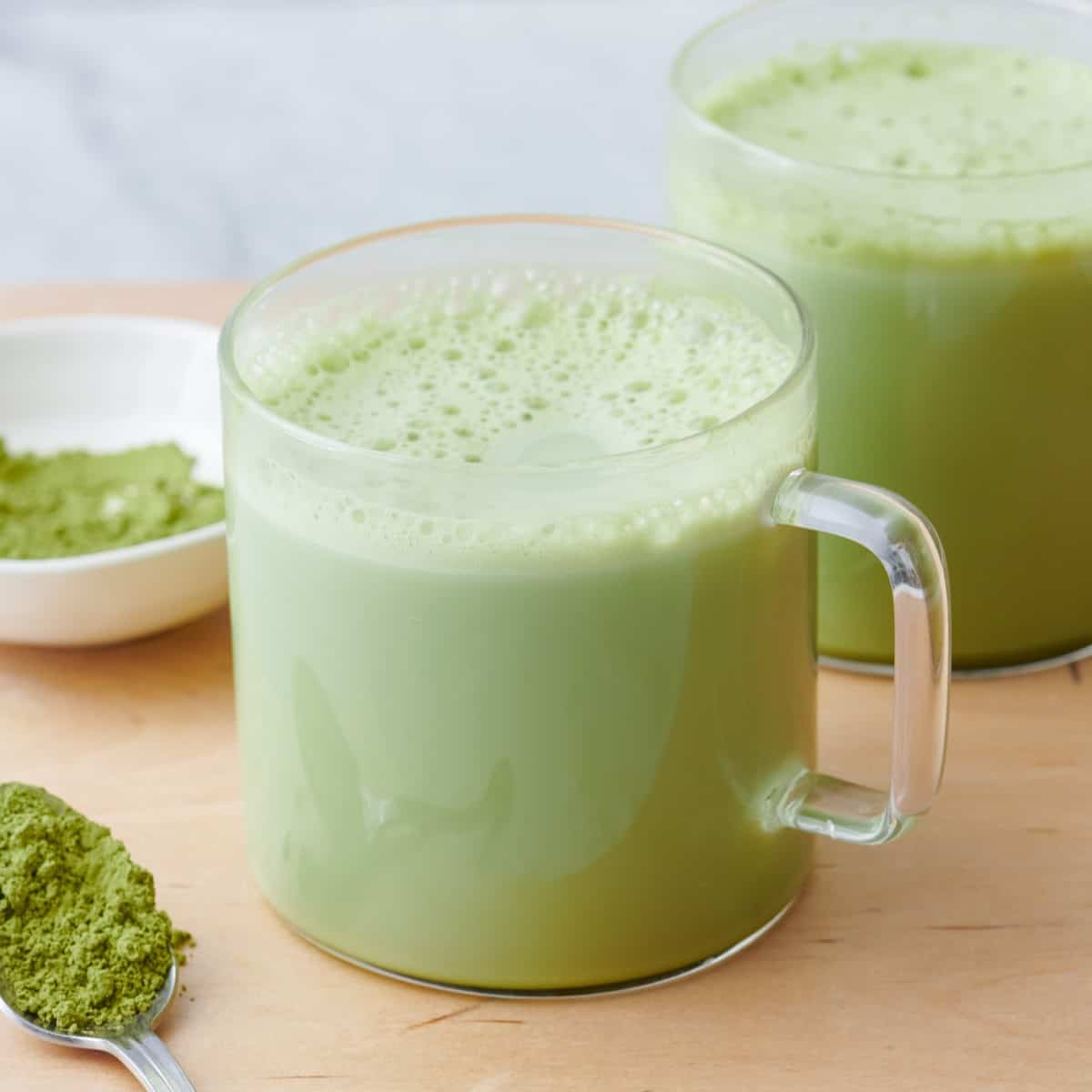 Matcha latte in a glass mug with matcha powder on a spoon and small dish.