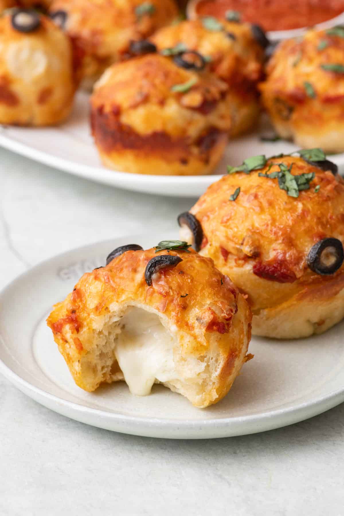 Pizza muffins on a plate with a bite taken out to show melty cheese with a plate of more nearby.