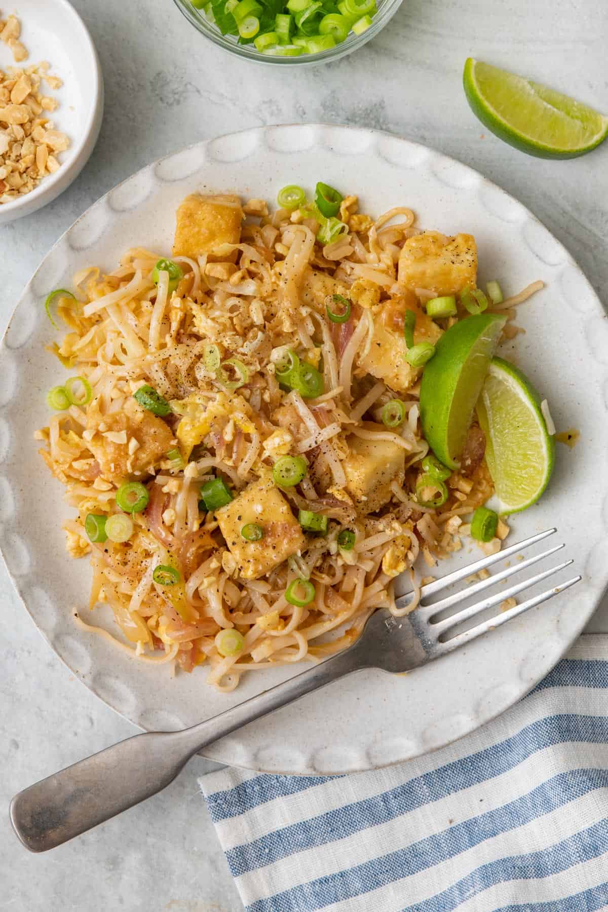 Vegetarian pad Thai with tofu cubes on a plate, garnished with green onions and lemon wedges.