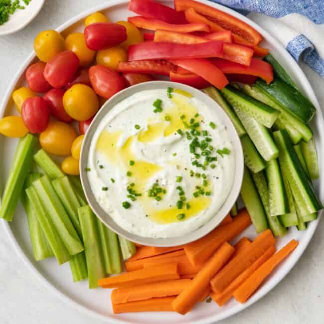 Whipped cottage cheese in a bowl garnished with chives and olive oil. Bowl on a platter surrounded by fresh veggies cut into sticks, including red bell peppers, zucchini, carrots, celery, and tomatoes.