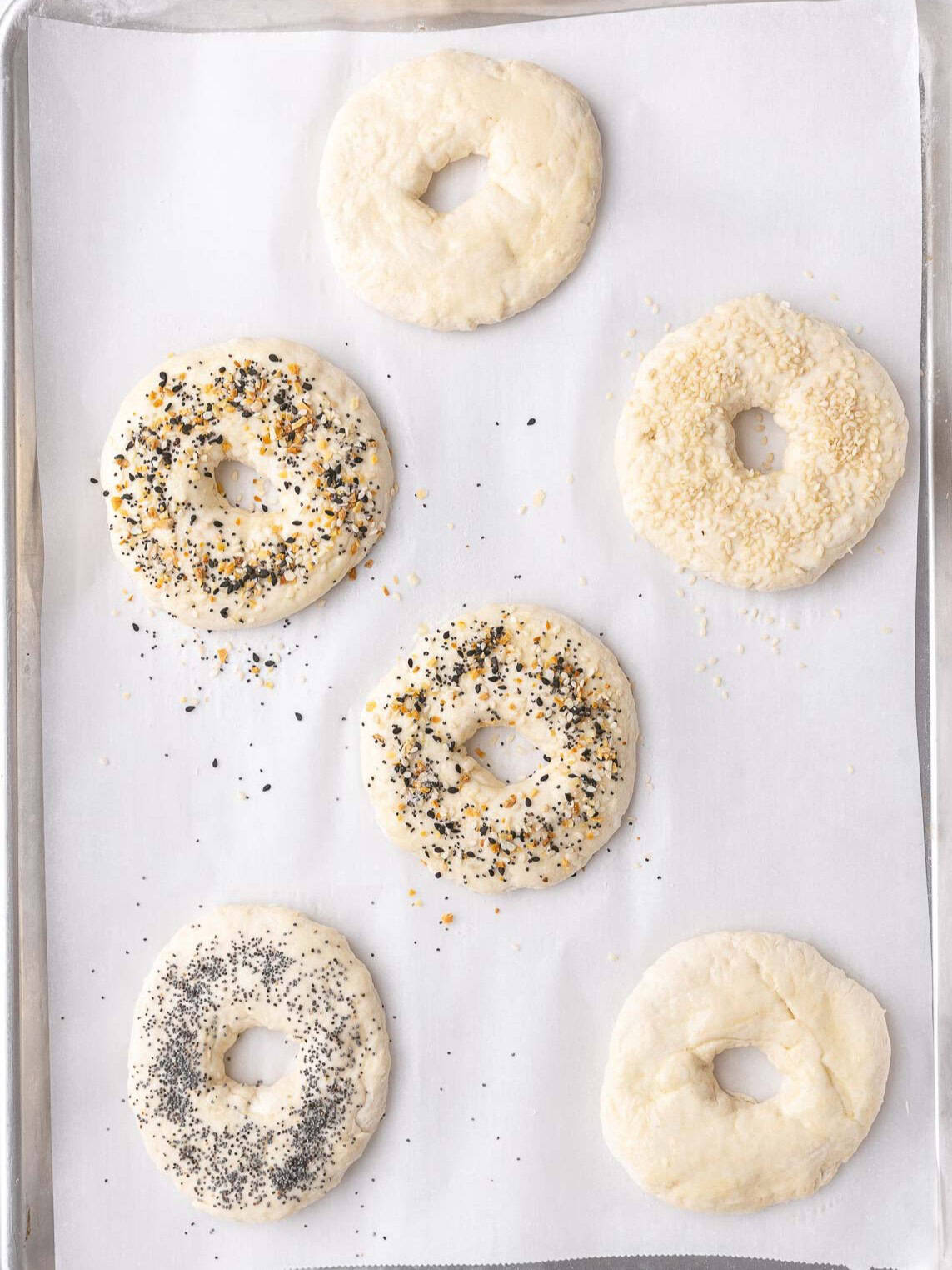 Bagels on a parchment lined baking sheet with egg white and toppings added before baking.