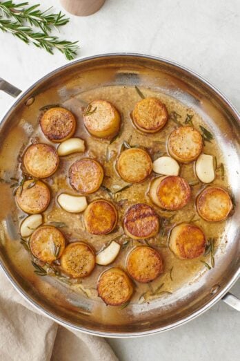 Fondant melting potatoes in a skillet with a butter sauce, rosemary, and garlic cloves.