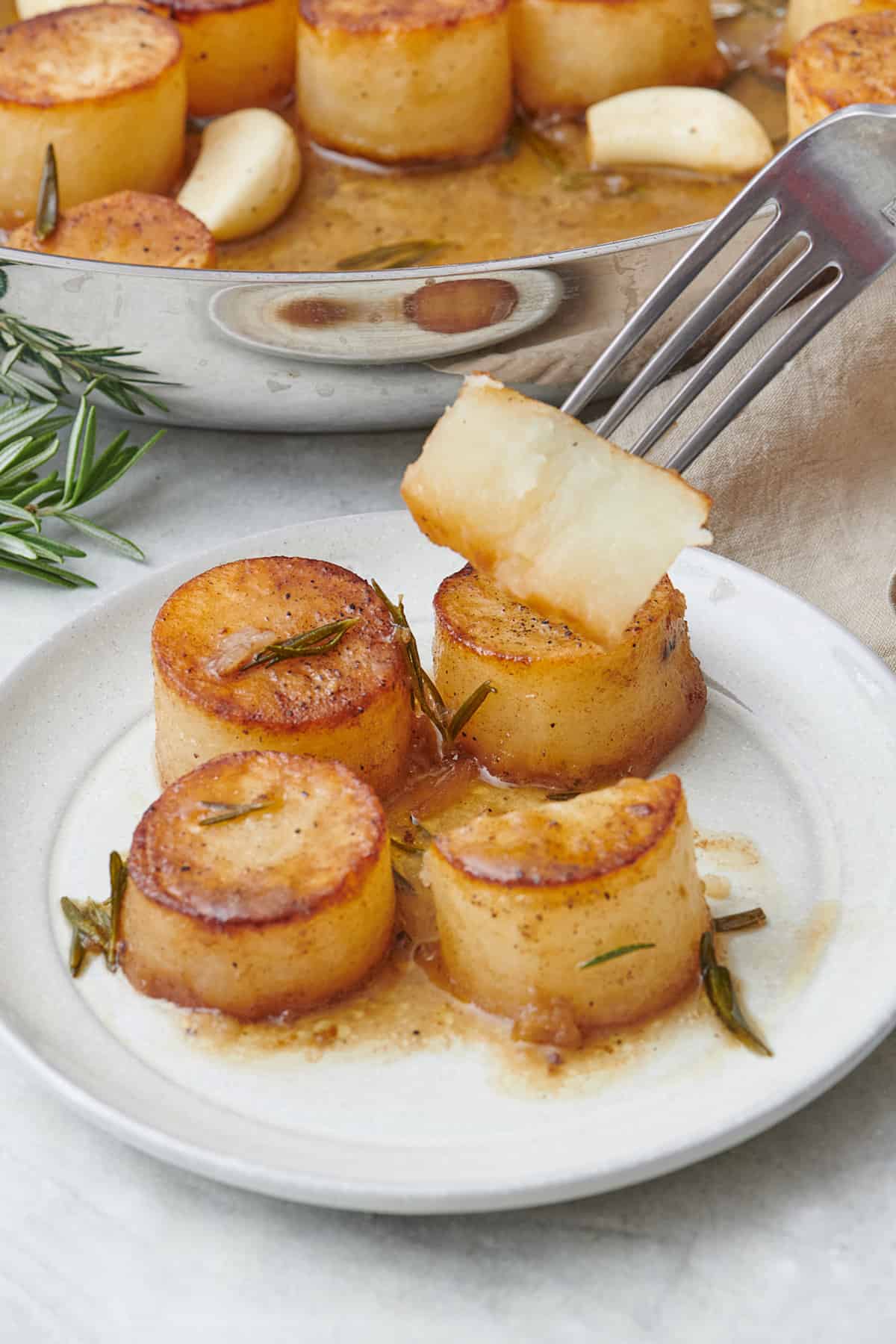 Fondant melting potatoes on a plate with a fork lifting up a piece to show inside texture. Skillet of more nearby.