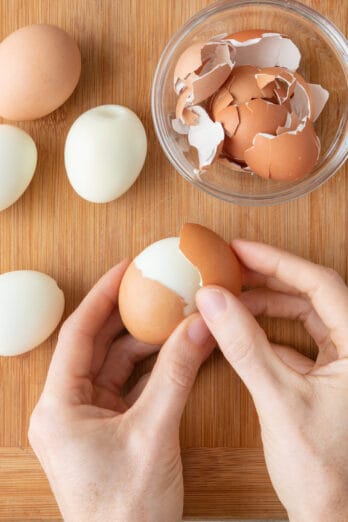 Hand peeling away the shell of a hard boiled egg with a small bowl of shell nearby.