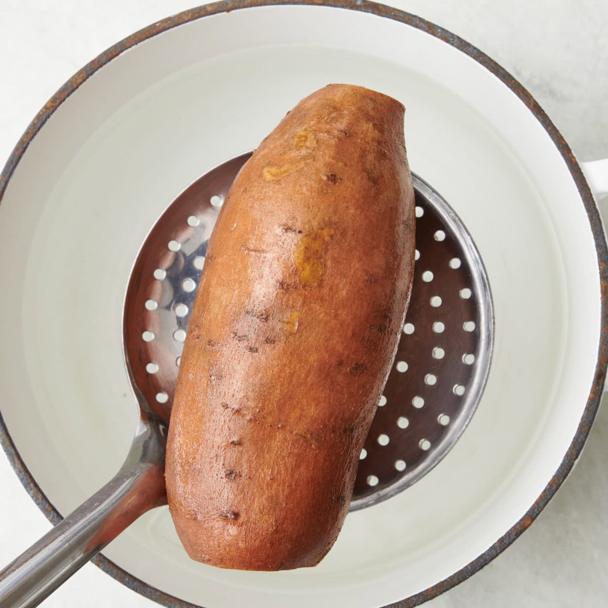 Whole sweet potato with root ends trimmed in a slotted spoon going into a pot of water before cooking.