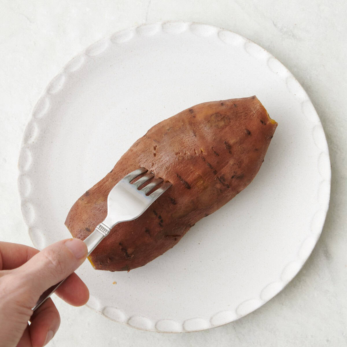 Sweet potato on a small plate with fork piercing whole sweet potato to show doneness.