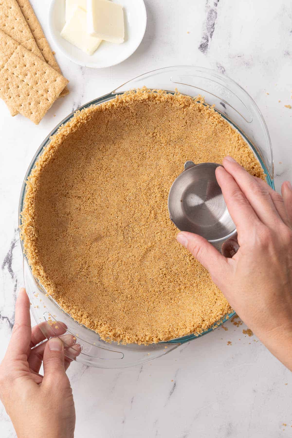 Tutorial showing how to make a graham cracker crust in a glass pie dish by pressing the crumbs down with a measuring cup.