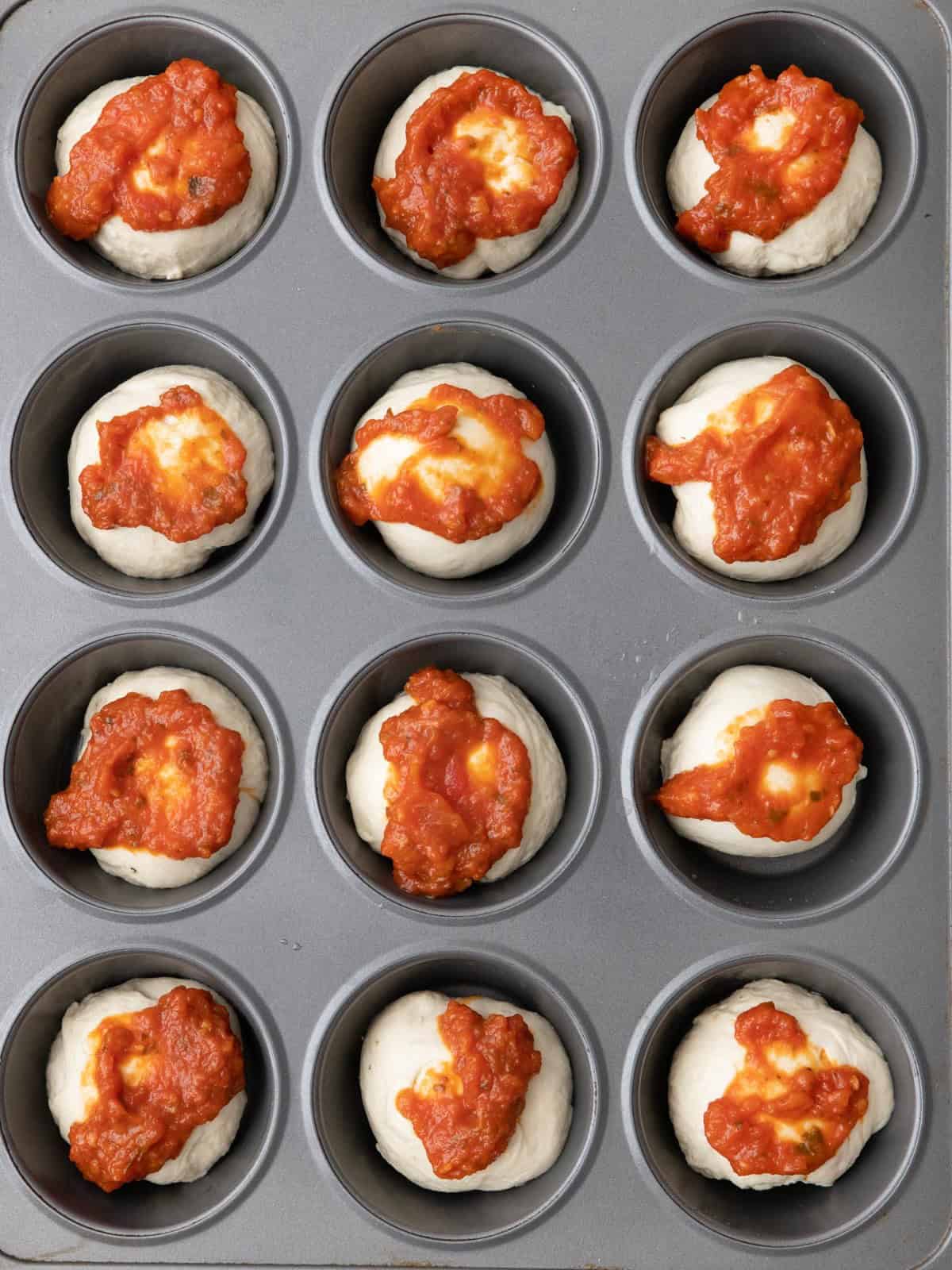Dough balls inside a muffin tin with pizza sauce on top.