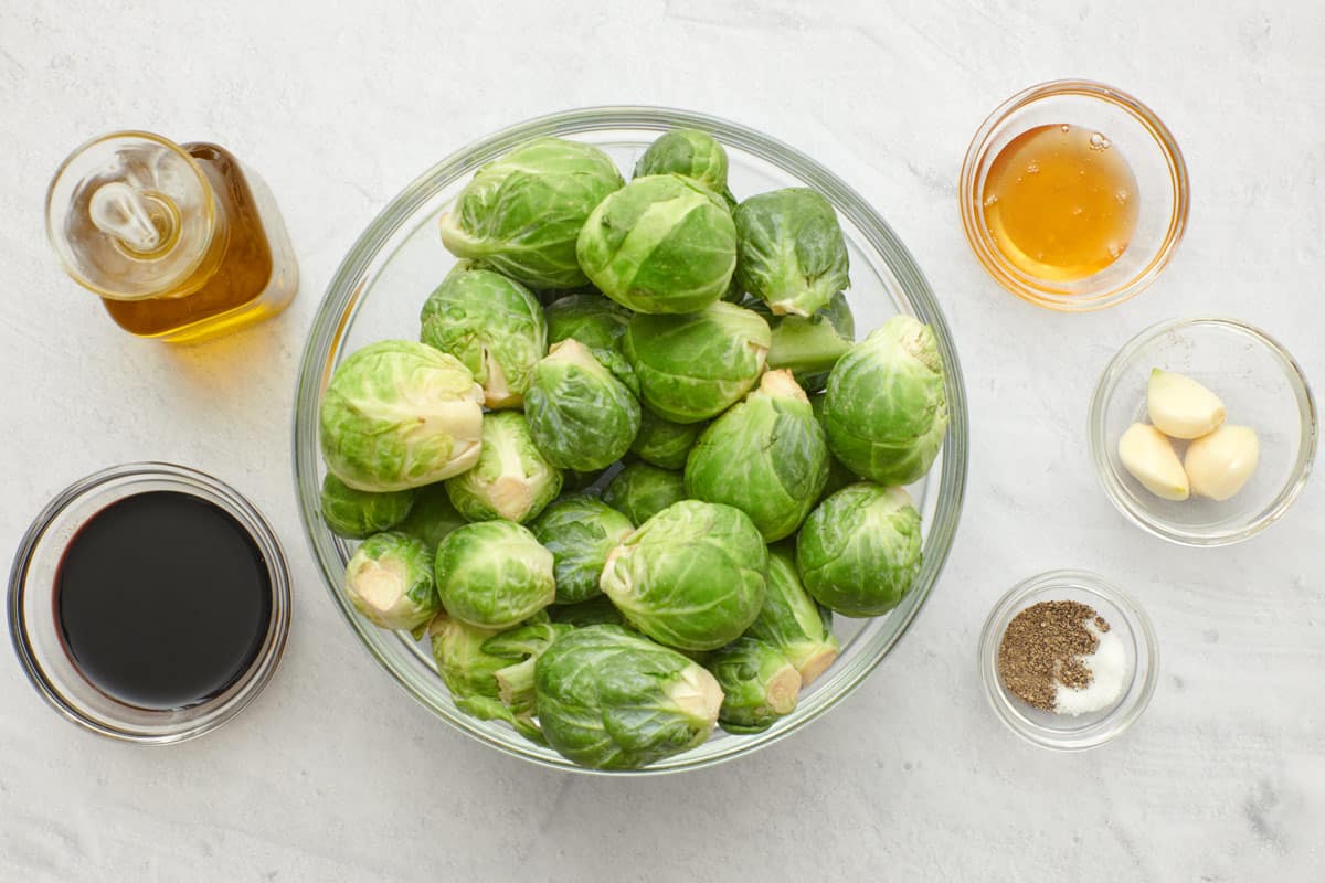 Ingredients before prepping in individual bowls and containers: oil, balsamic vinegar, fresh brussel sprouts, honey, garlic cloves, salt and pepper.