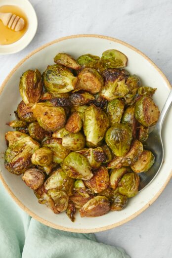 Balsamic roasted brussel sprouts in a serving bowl with spoon dipped in and small dish of honey nearby.