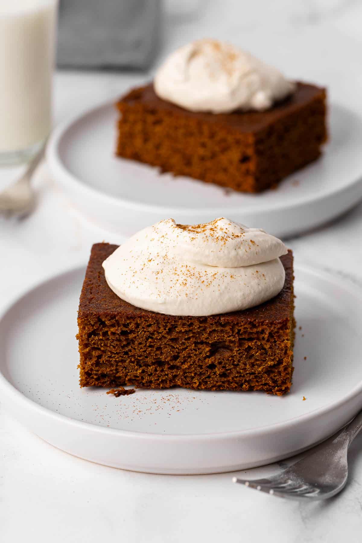 2 small plates with a slice of gingerbread topped with soft whipped cream and ground cinnamon.