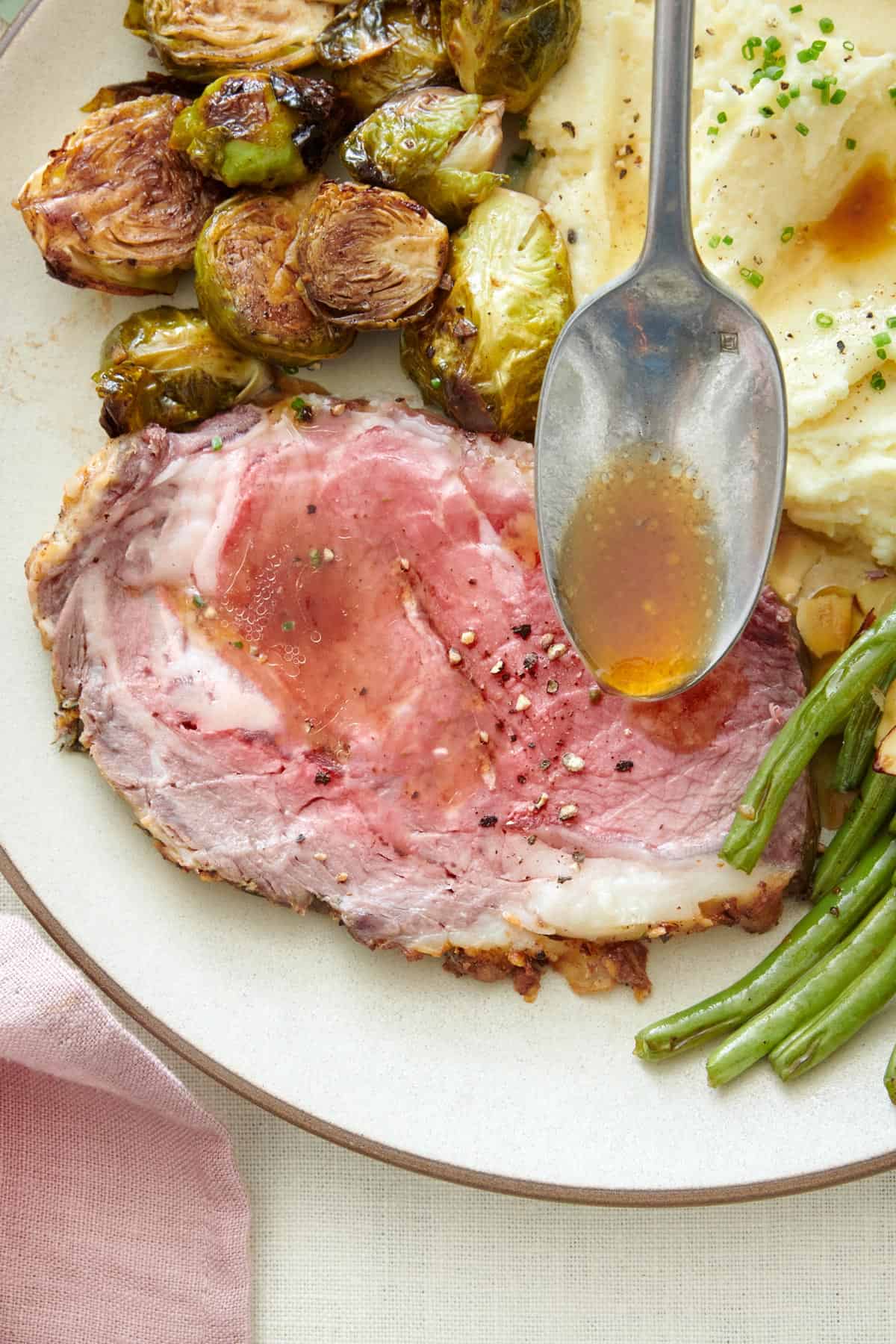 Spoon pouring au jus over a slice of prime rib on a plate next to brussel sprouts, mashed potatoes, and green beans.