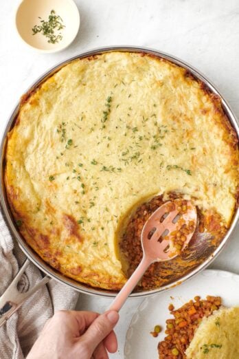Lentil shepherd’s pie garnished with fresh thyme and a serving removed on a plate nearby, serving spoon dipped into skillet.