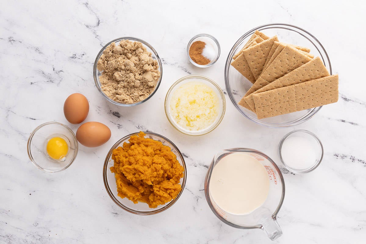 Ingredients to make recipe in individual bowls and containers: egg yolk plus 2 whole eggs, brown sugar, pumpkin puree, melted butter, spices and salt, graham cracker sheets, milk, and sugar.