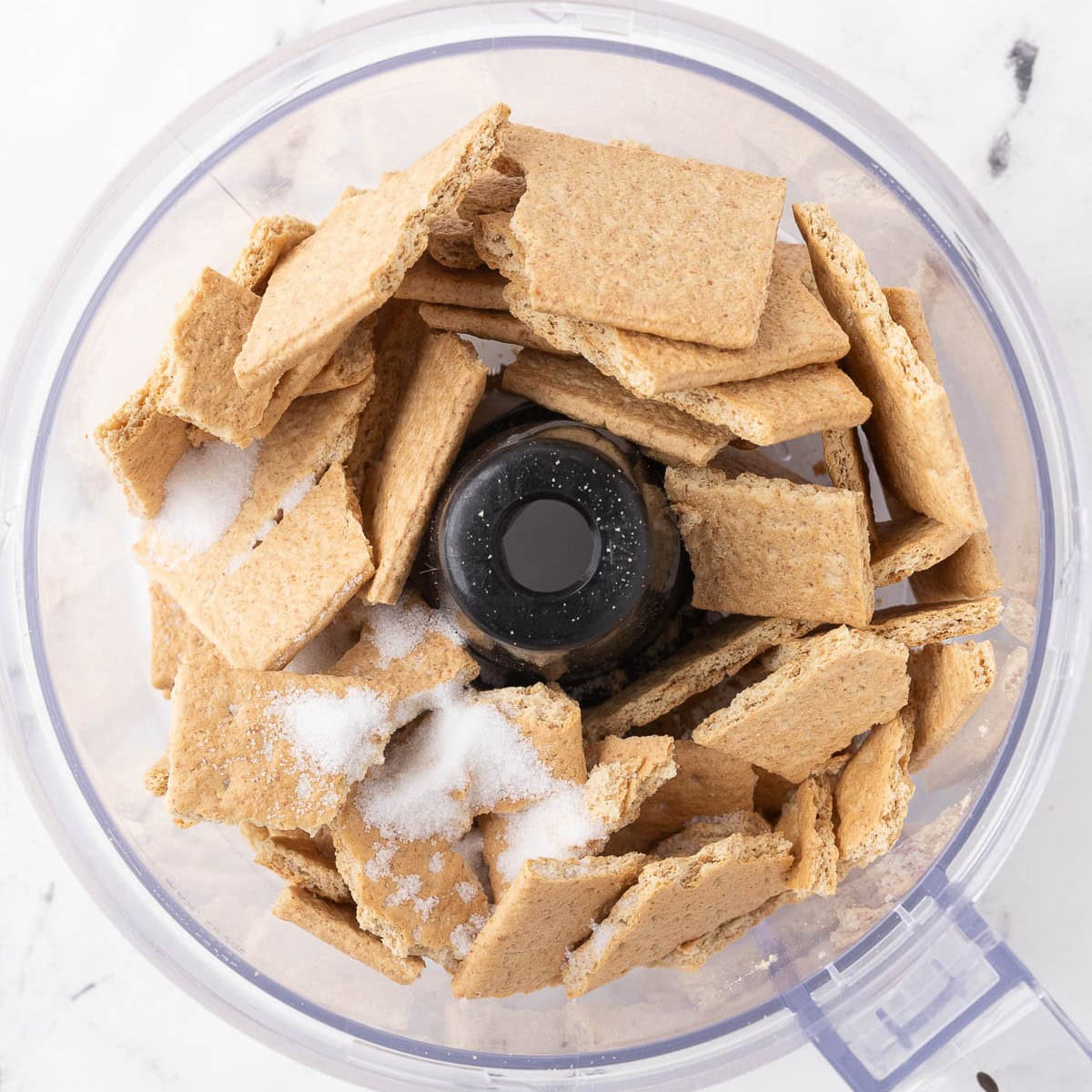 Graham crackers and sugar in the bowl of a food processor, before processing.
