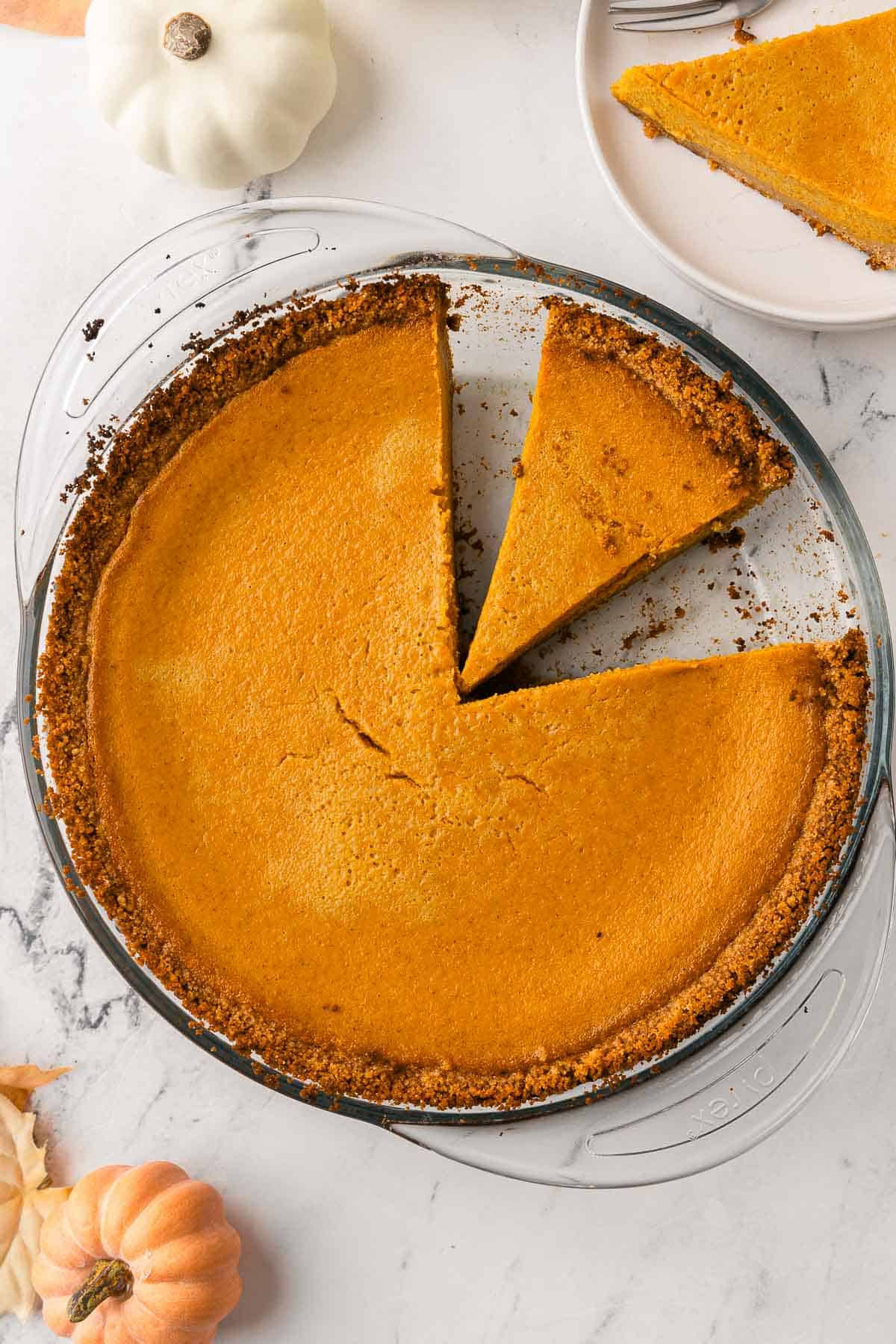 Pumpkin pie with a graham cracker crust. Two slices cut from the pie with one on a plate nearby.