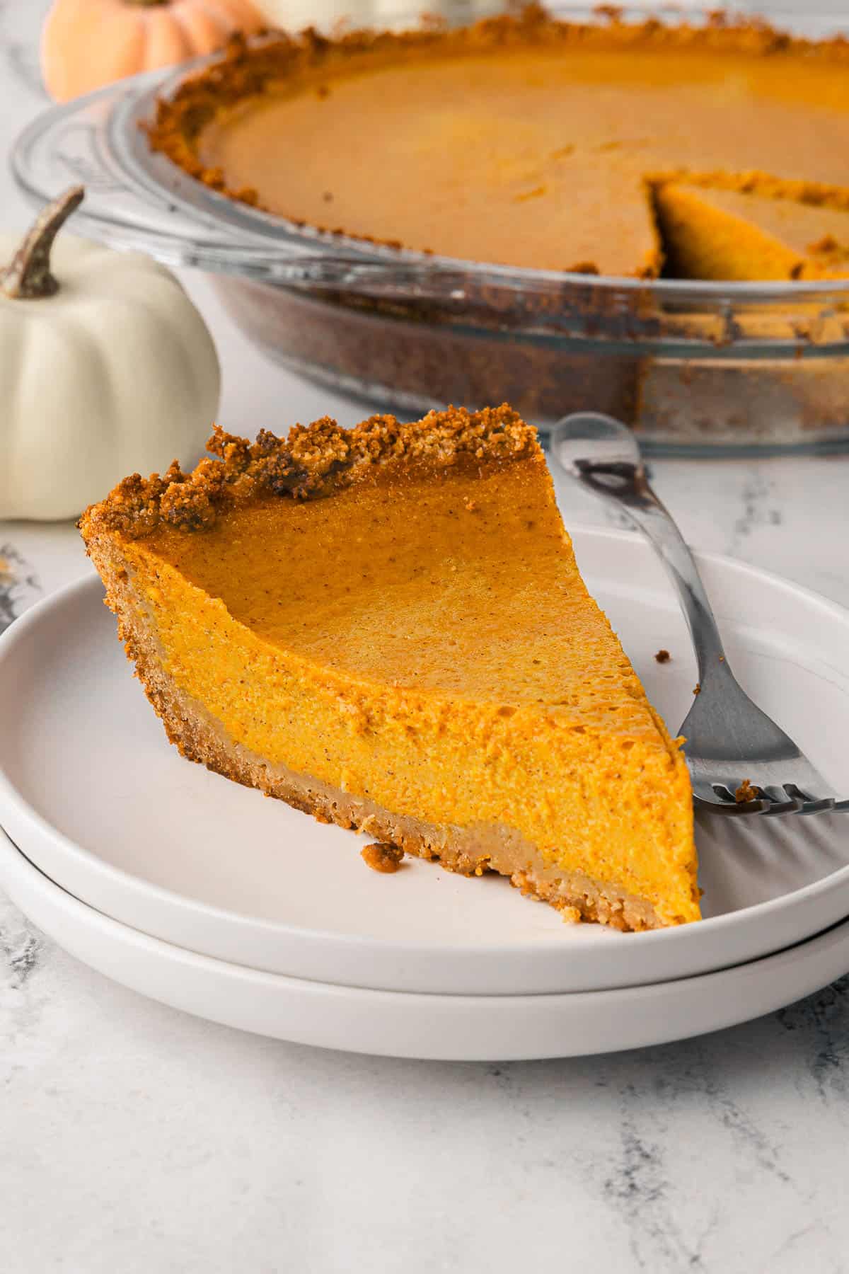 Slice of pumpkin pie on a plate with whole pie in a glass pie dish nearby.