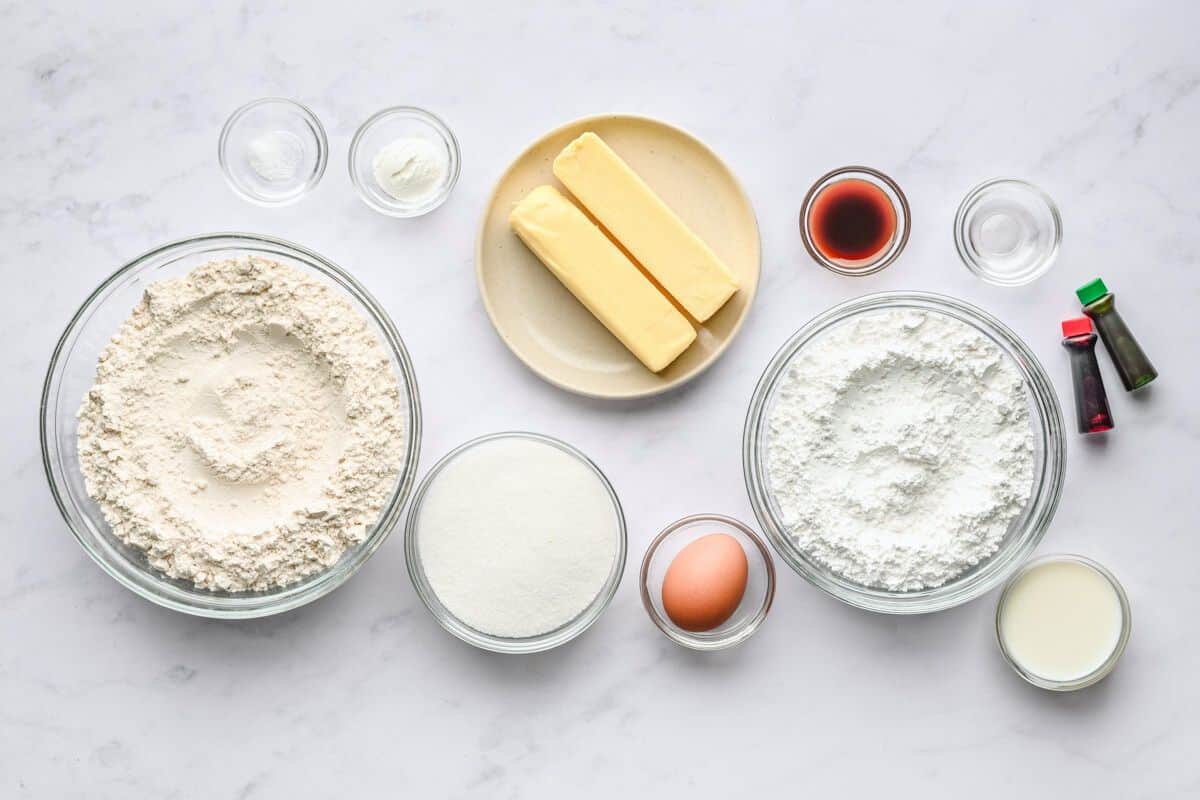 Ingredients for recipe in individual bowls and containers: flour, salt, baking powder, butter sticks, sugar, egg, vanilla, peppermint extract, powdered sugar, food coloring, and cream.