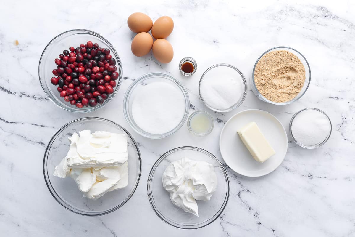 Ingredients for recipe in individual bowls: cranberries, cream cheese, eggs, sugar, sour cream, vanilla, lemon juice, butter, graham cracker crumbs.