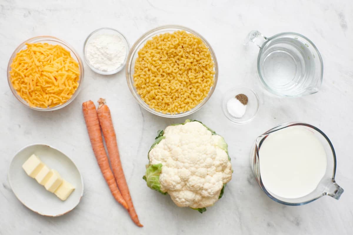 Ingredients for recipe in individual bowls: shredded cheese, butter, flour, elbow macaroni, carrots, cauliflower, salt and pepper, water, and milk.