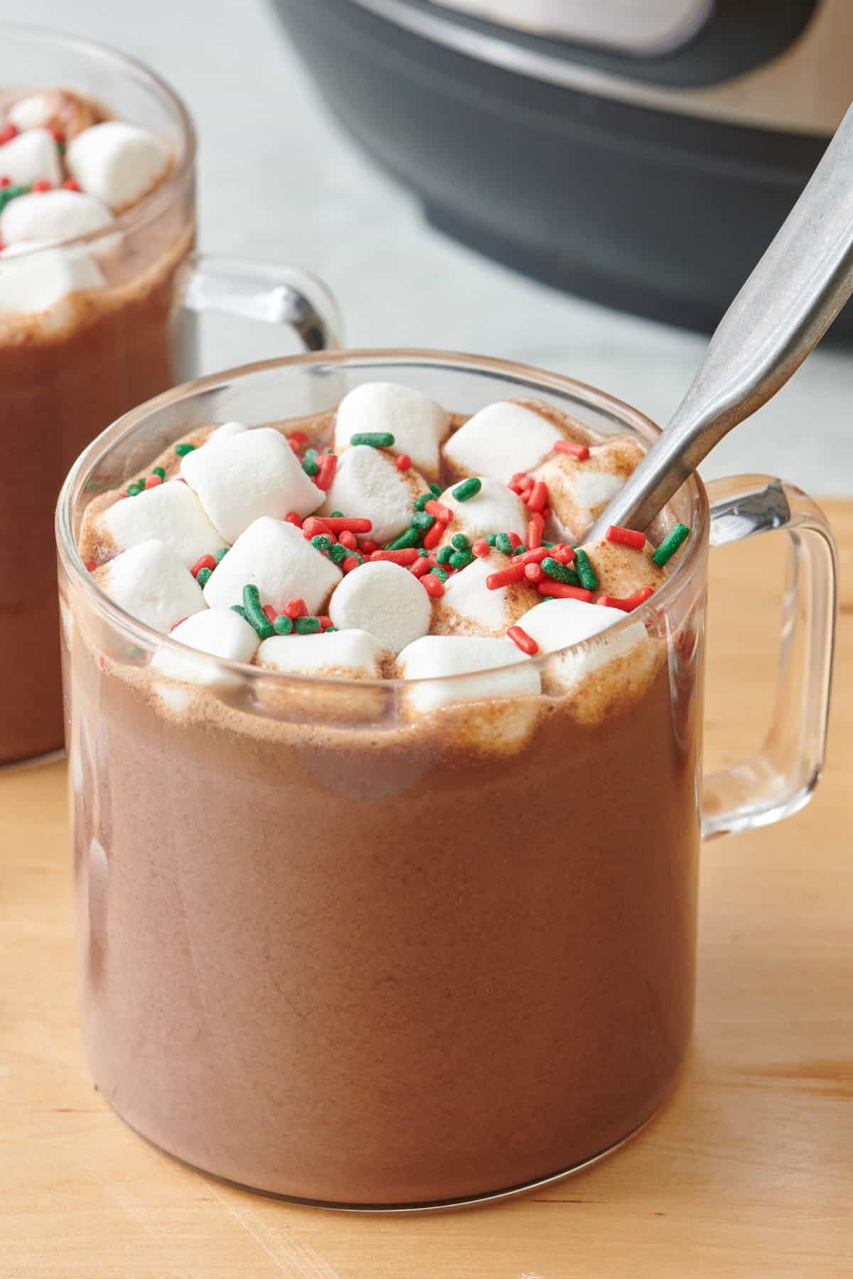 Homemade hot chocolate in a glass mug with a spoon dipped in and topped with mini marshmallows and sprinkles.
