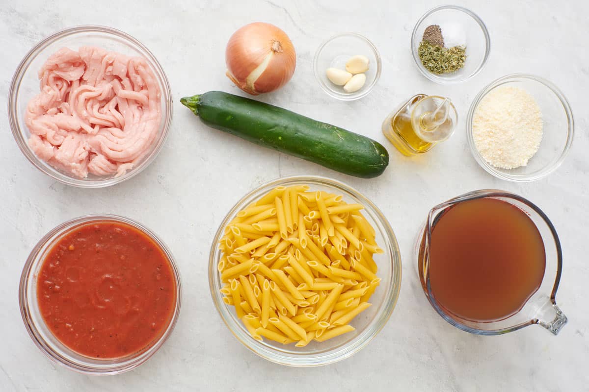 Ingredients for recipe in individual bowls before prepping: ground turkey, marinara sauce, onion, zucchini, dry penne pasta, garlic cloves, seasonings, oil, parmesan, and broth.