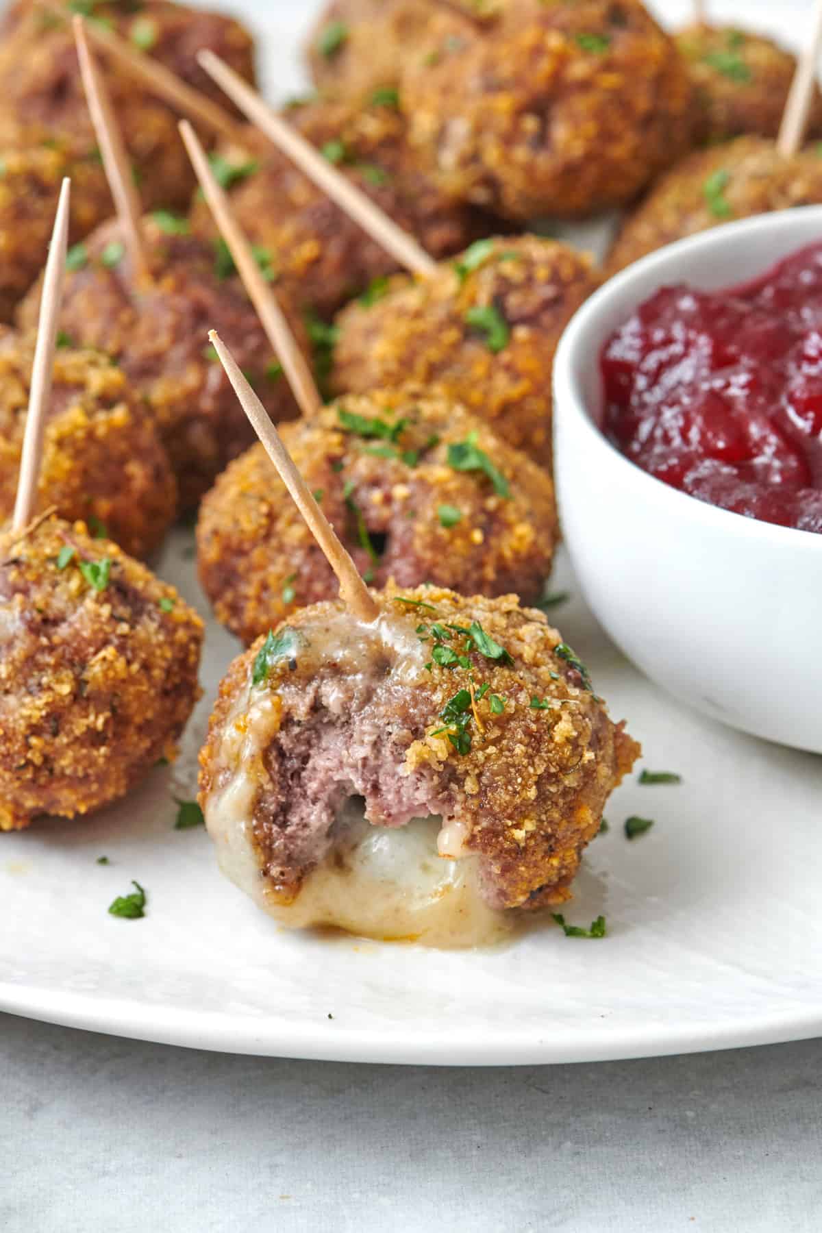 Close up of one brie cheese stuffed meatball with a bite taken out to show the melty cheese coming out. More meatballs and cranberry sauce around.