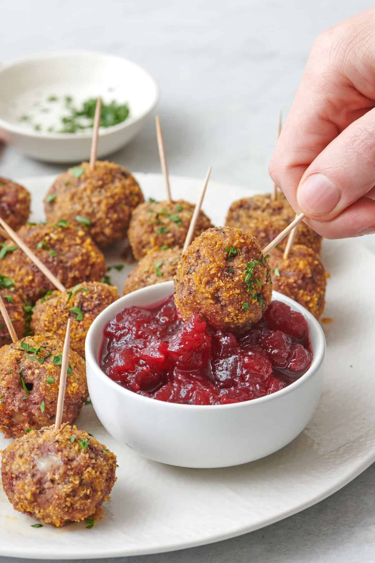 Hand dipping a crispy breadcrumb coated stuffed meatball in a small dish of cranberry sauce on a plate with more meatballs.
