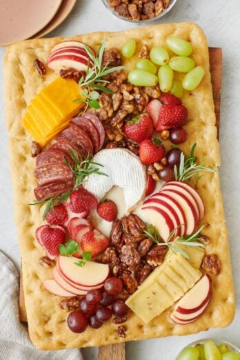 Focaccia bread with baked brie in the center served as a charcuterie board with fresh fruit, nuts, cheeses, and deli meat.