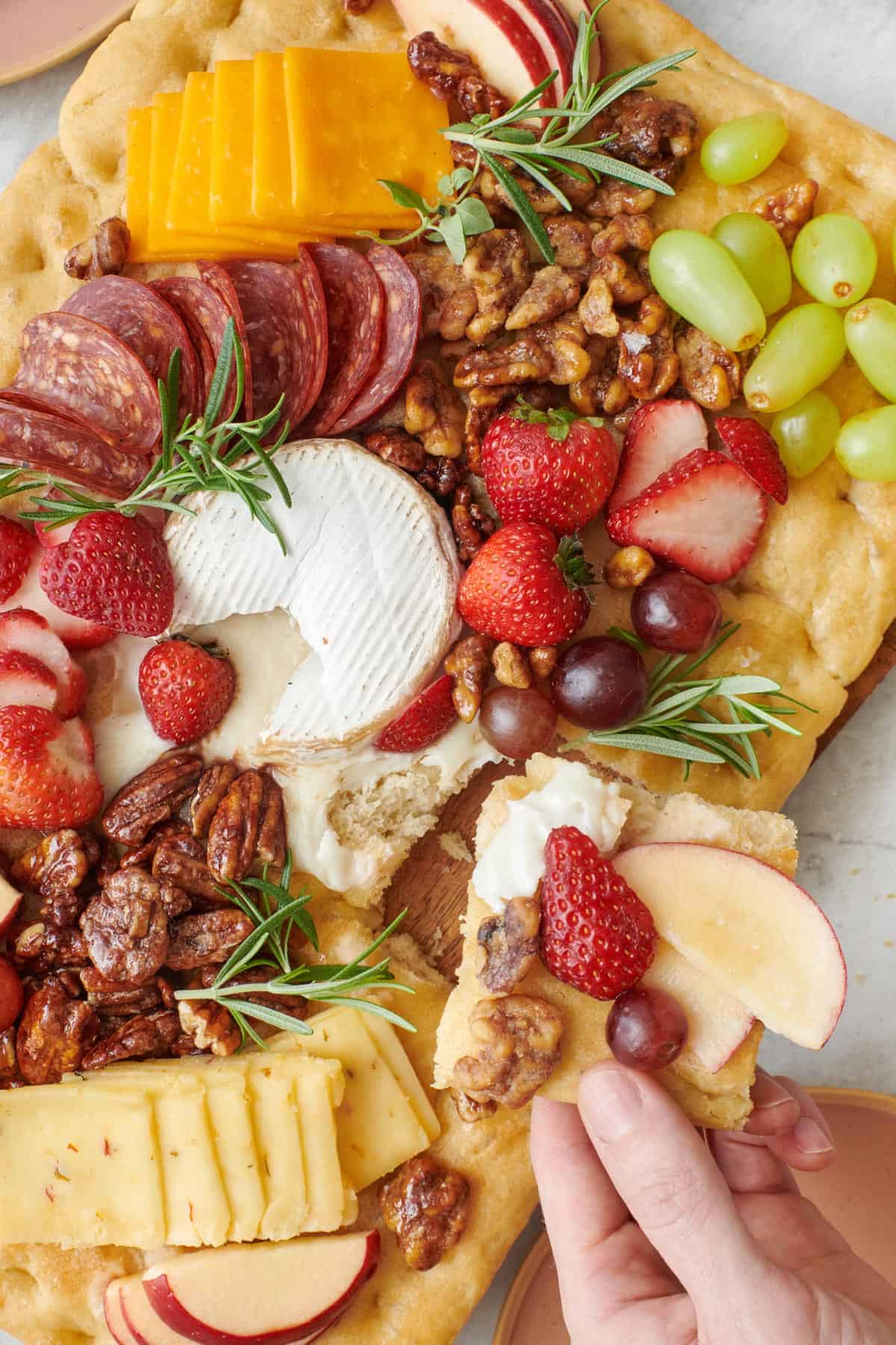 Baked brie focaccia charcuterie board filled with fresh fruit, nuts, cheeses, and deli meat with a hand lifting up a chip with some on it.