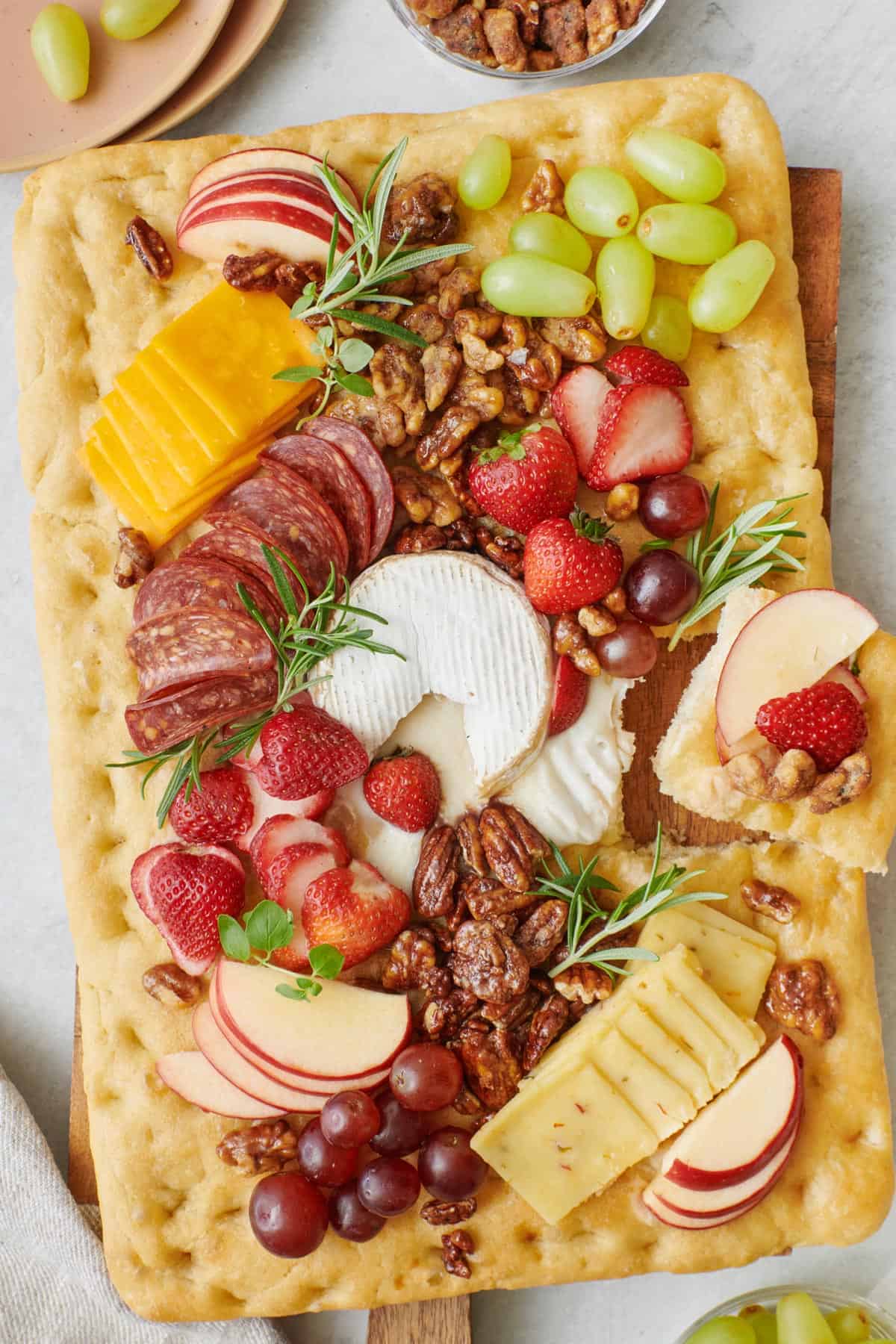 Focaccia charcuterie board with melty baked brie, covered with fresh fruit, nuts, cheeses, and deli meat with a hand lifting up a chip with some on it.