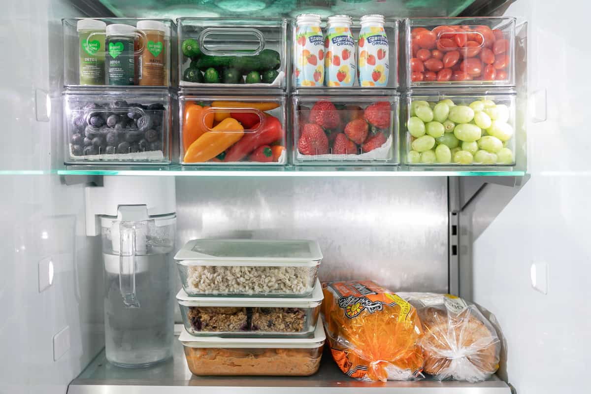 Bread, leftovers, and water stocked on the bottom shelf of a fridge with fresh produce and small individual foods like yogurt in containers.
