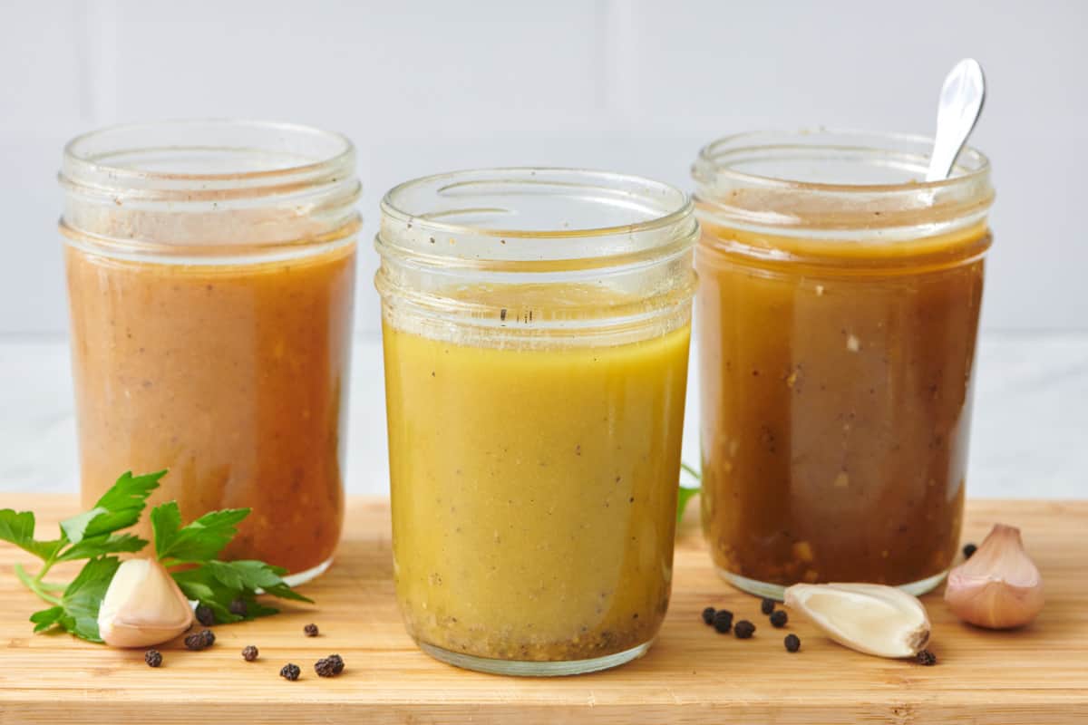 Three different flavors of homemade vinaigrette dressing in small mason jars. Extra ingredients like garlic cloves and peppercorns around.