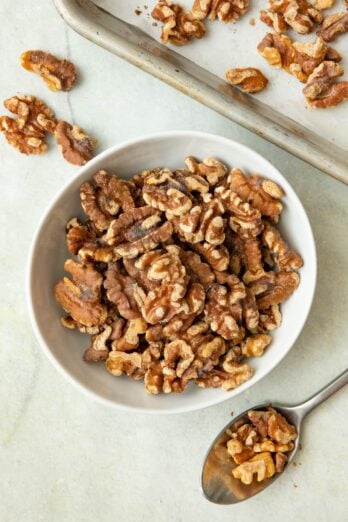 Toasted walnuts in a bowl with sheet pan and spoon nearby.