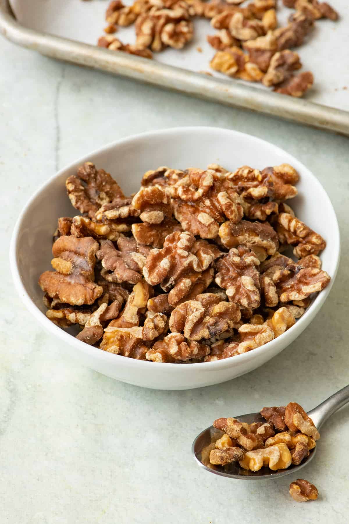 Bowl of toasted walnuts with a spoon of scooped walnuts nearby and a sheet pan behind the bowl.