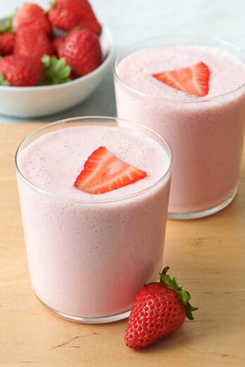 Two glasses of strawberry yogurt smoothie with a slice of strawberry on top and a small dish of more fresh strawberries nearby.