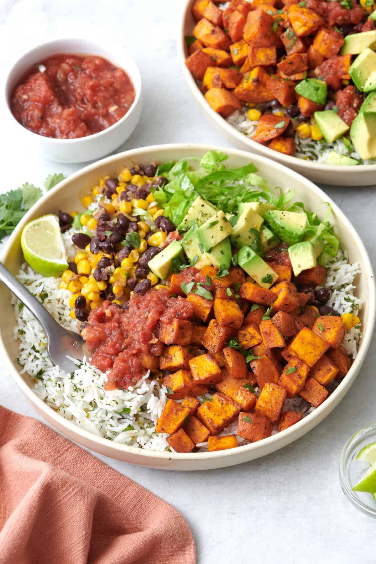 Two vegetarian burrito bowls made with sweet potatoes, black bean and corn, cilantro lime rice, lettuce, avocado, salsa, and lime wedges.