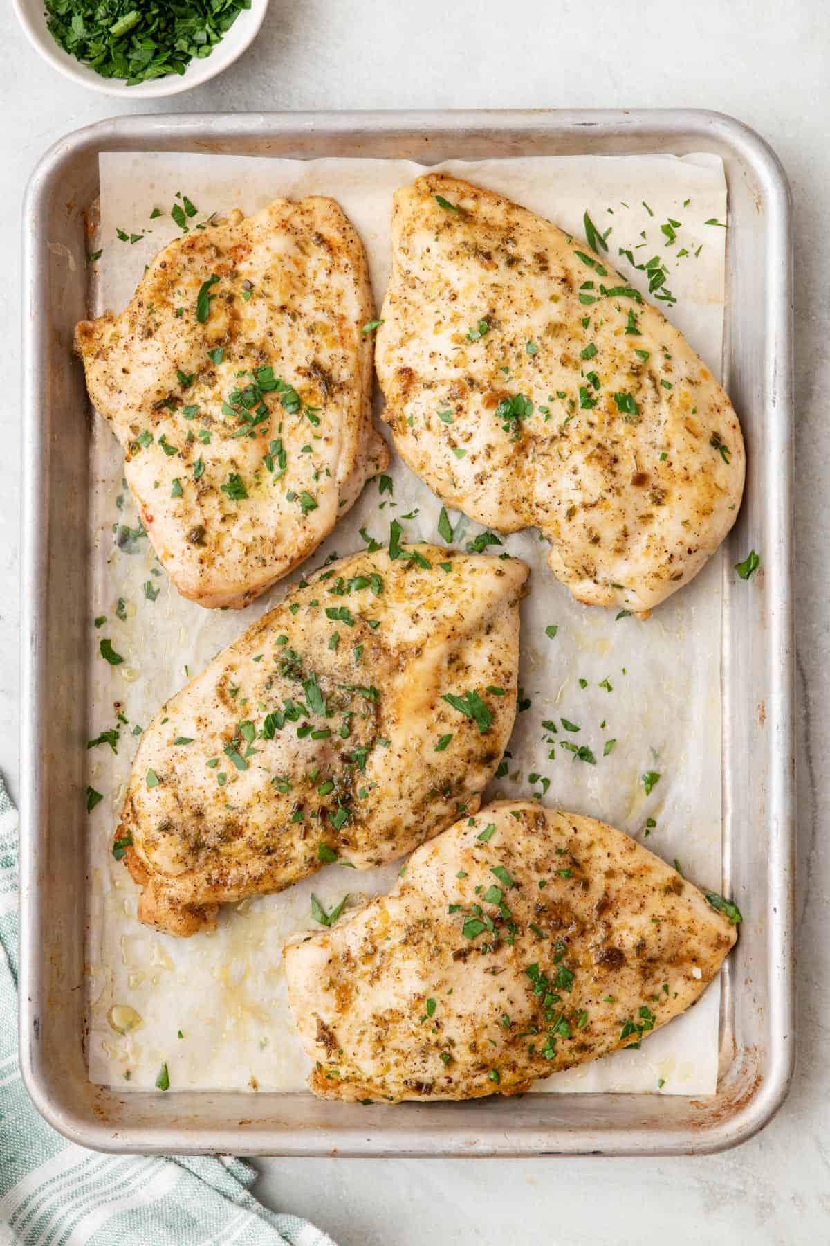 A baking sheet with 4 baked ranch chicken breasts garnished with parsley on parchment paper.