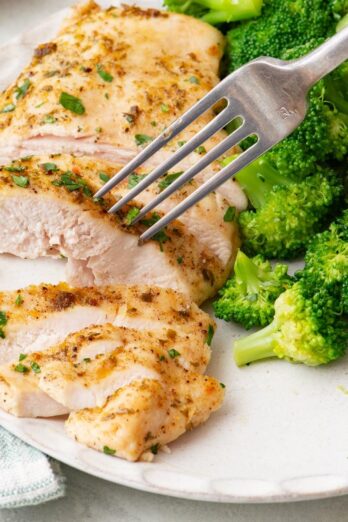 Close-up of sliced baked ranch chicken being stabbed by a fork on a white plate with a side of broccoli.