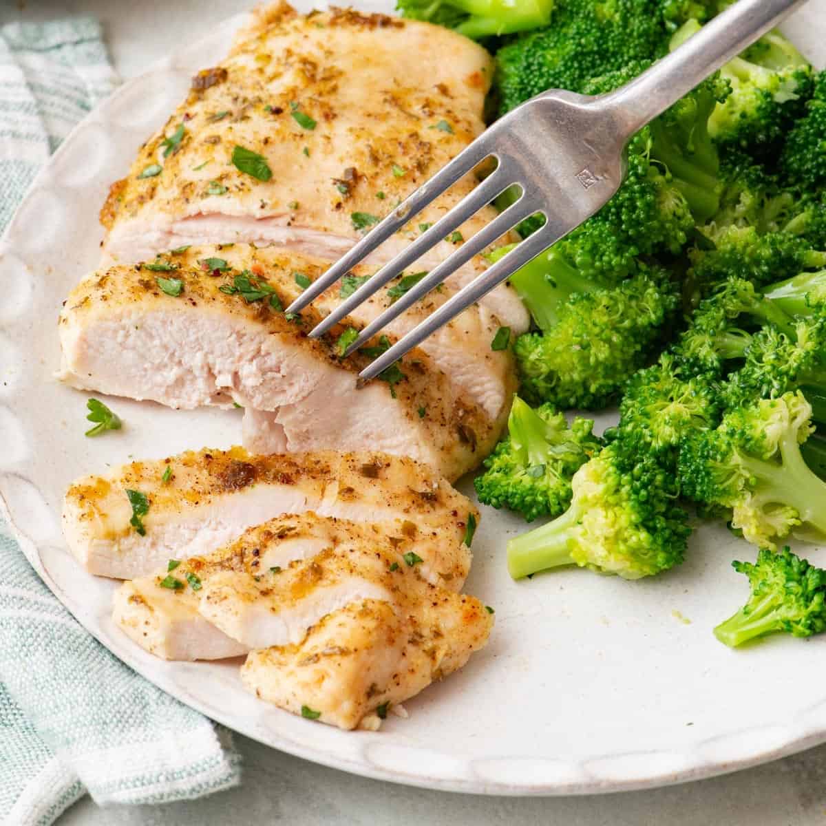 Close-up of sliced baked ranch chicken being stabbed by a fork on a white plate with a side of broccoli.