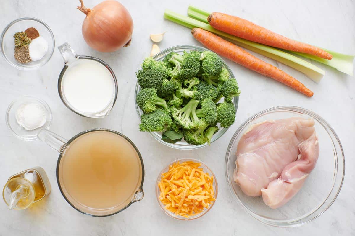 Ingredients for recipe in individual bowls: spices, flour, oil, onion, cream, broth, garlic cloves, broccoli florets, shredded cheddar cheese, carrots and celery, and chicken breasts.