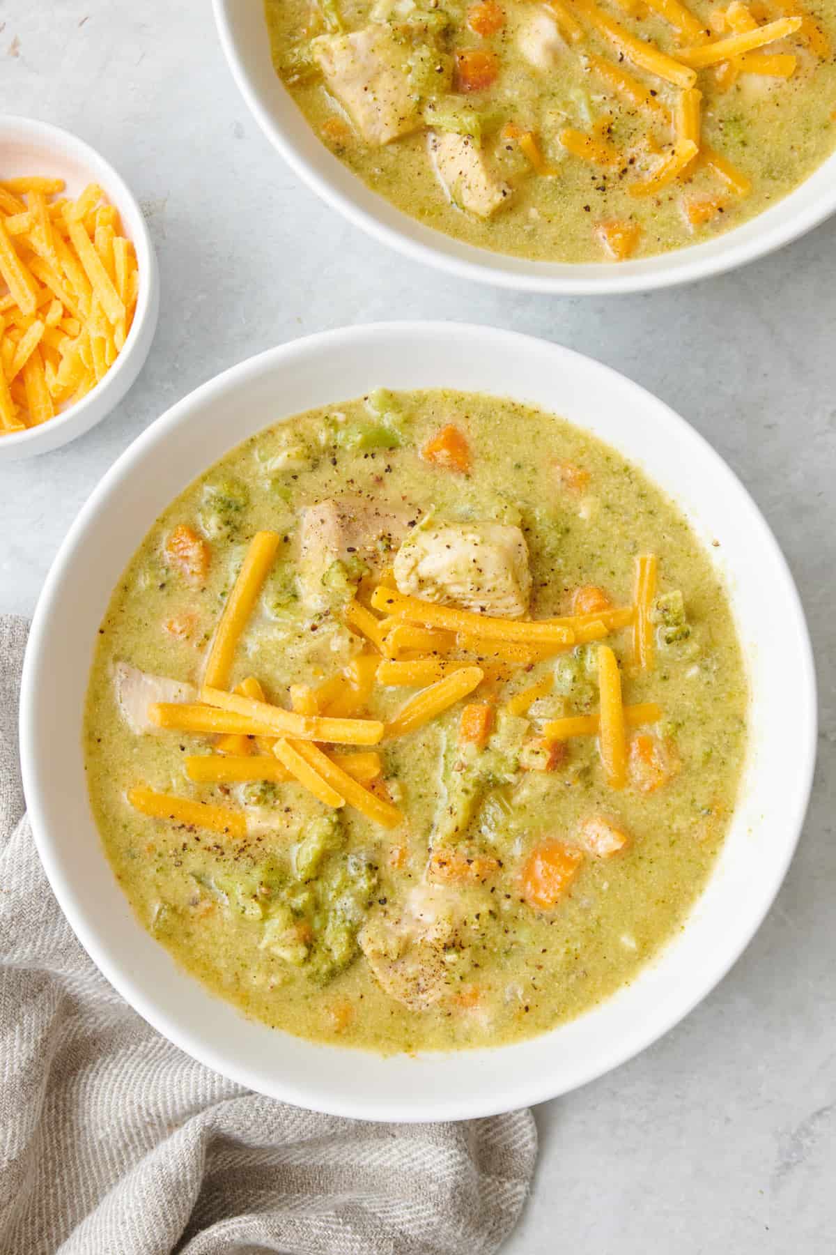 Two bowls of creamy chicken broccoli soup with shredded cheddar cheese on top.