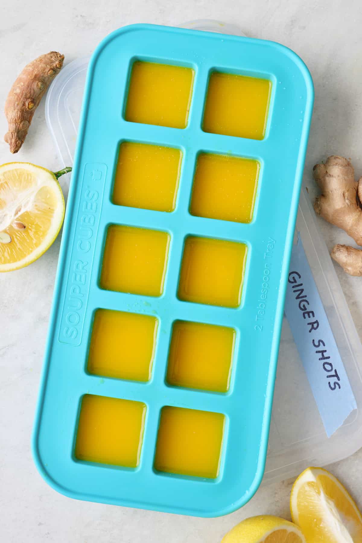 Ginger shots in an ice cube tray with lemons and ginger roots nearby.