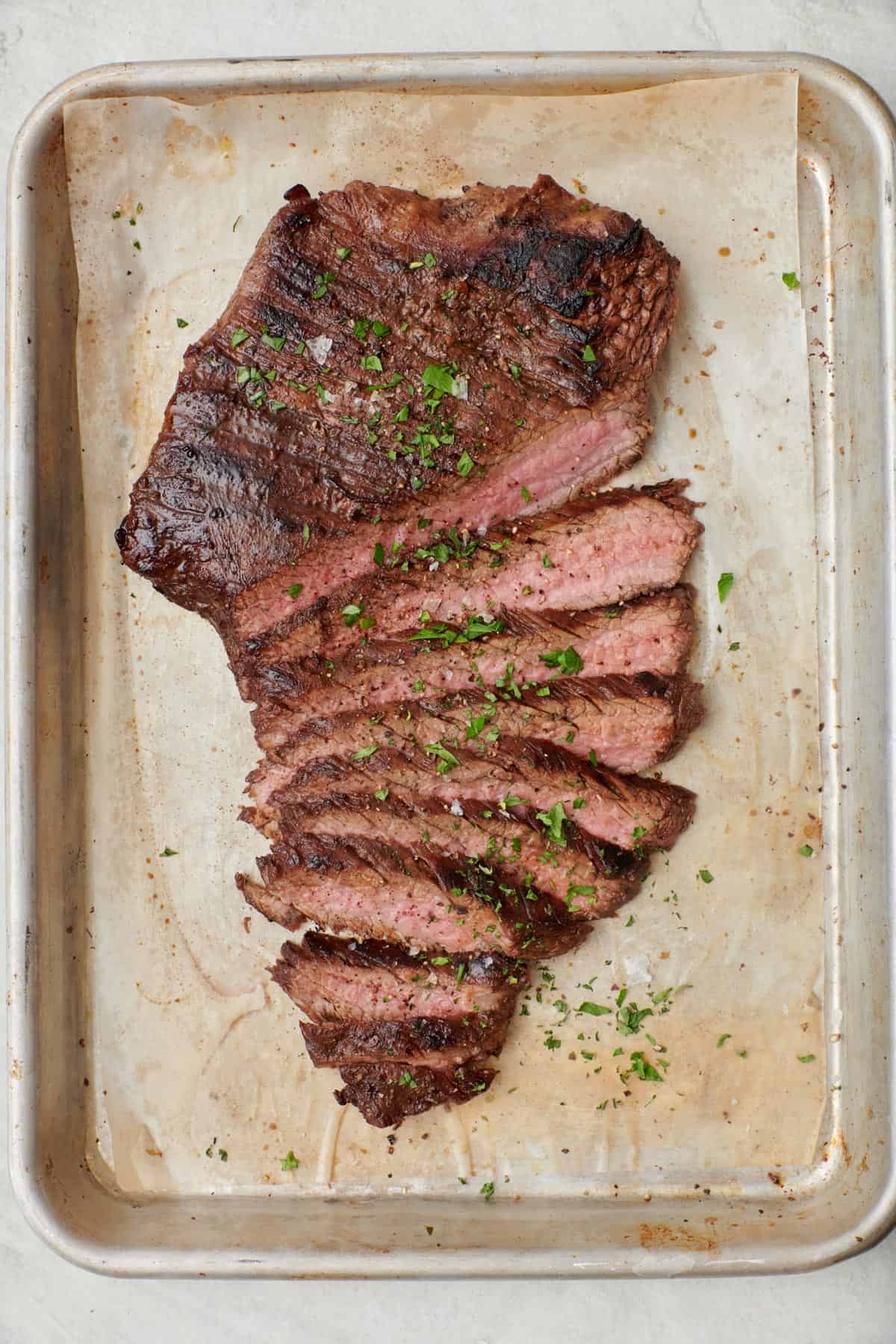 Flank steak on a parchment-lined sheet pan after cooking in the oven, partially sliced and cooked to to rare.