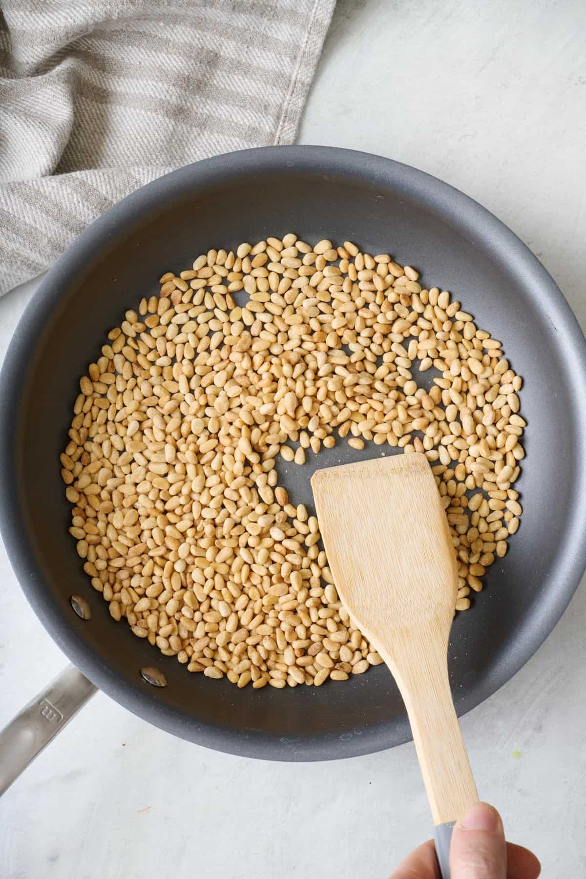 Toasted pine nuts in a small skillet with a wooden spatula pushing them around.