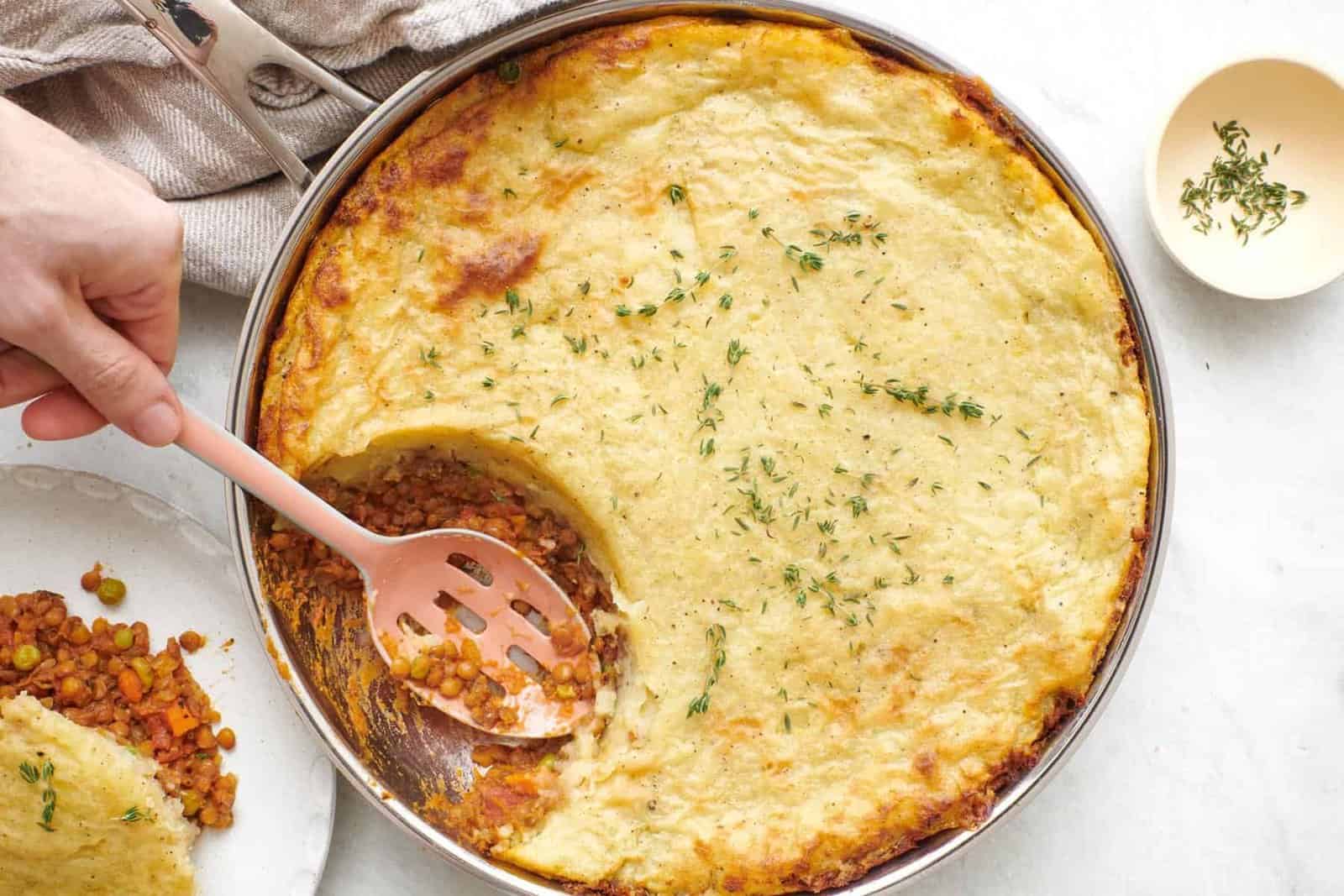 Lentil shepherd’s pie garnished with fresh thyme and a serving removed on a plate nearby, serving spoon dipped into skillet.