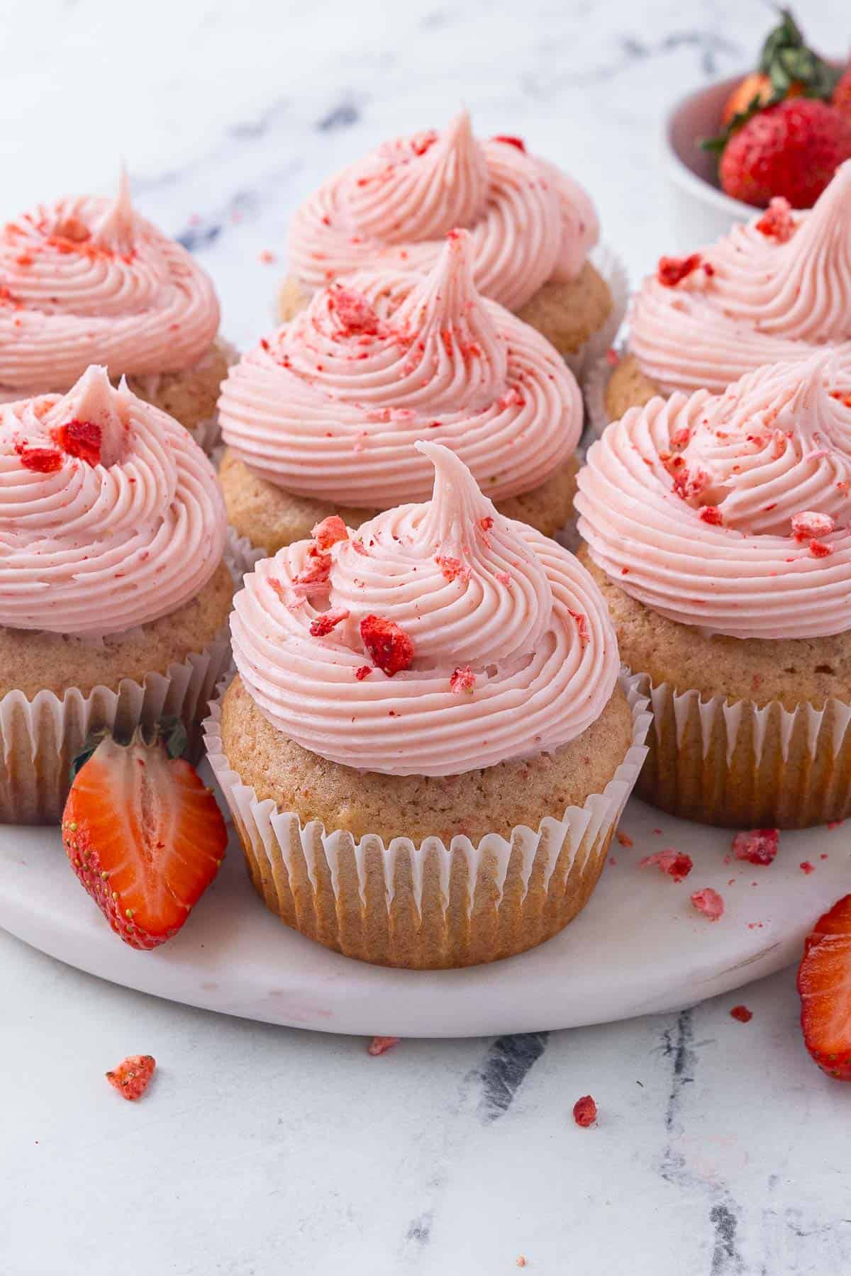 Strawberry cupcakes in liners topped with pink strawberry cream cheese on a round marble serving plate.