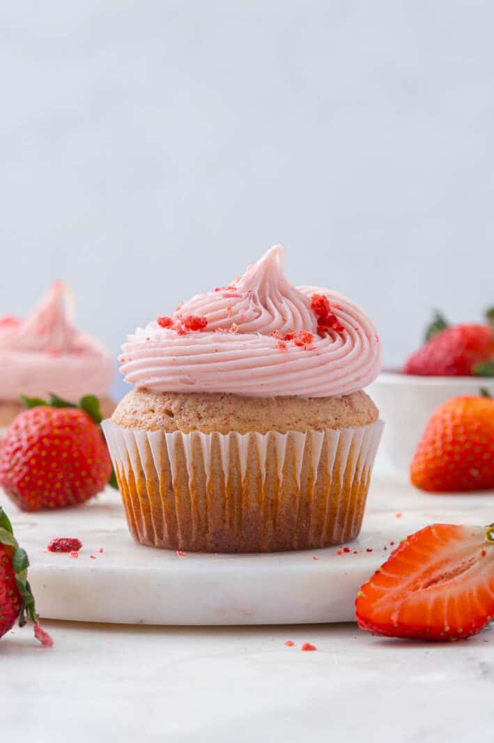 Close up of a strawberry cupcake with pink strawberry cream cheese frosting surrounded by fresh strawberries