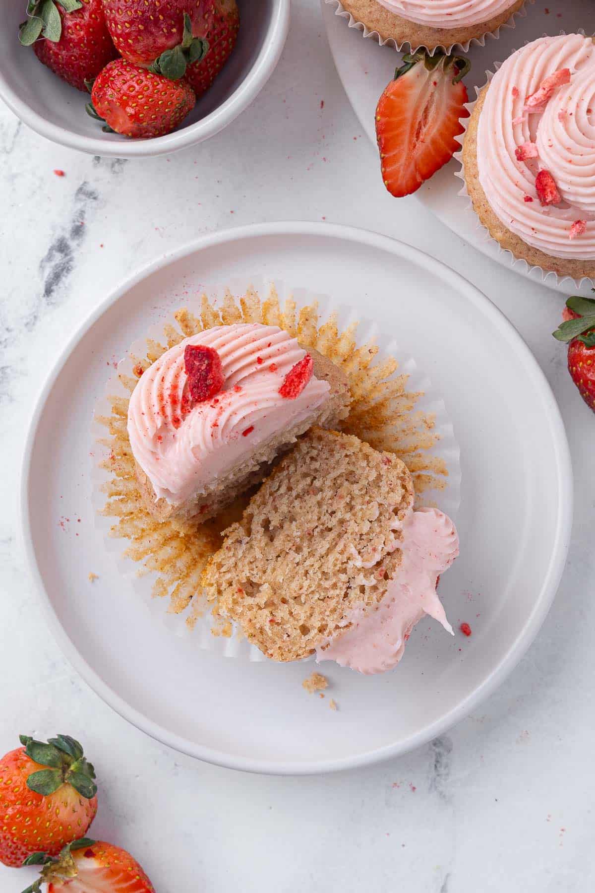 Overhead view of a strawberry cupcake cut open with one cut side facing upwards; plate of other cupcakes and bowl of fresh strawberries nearby.